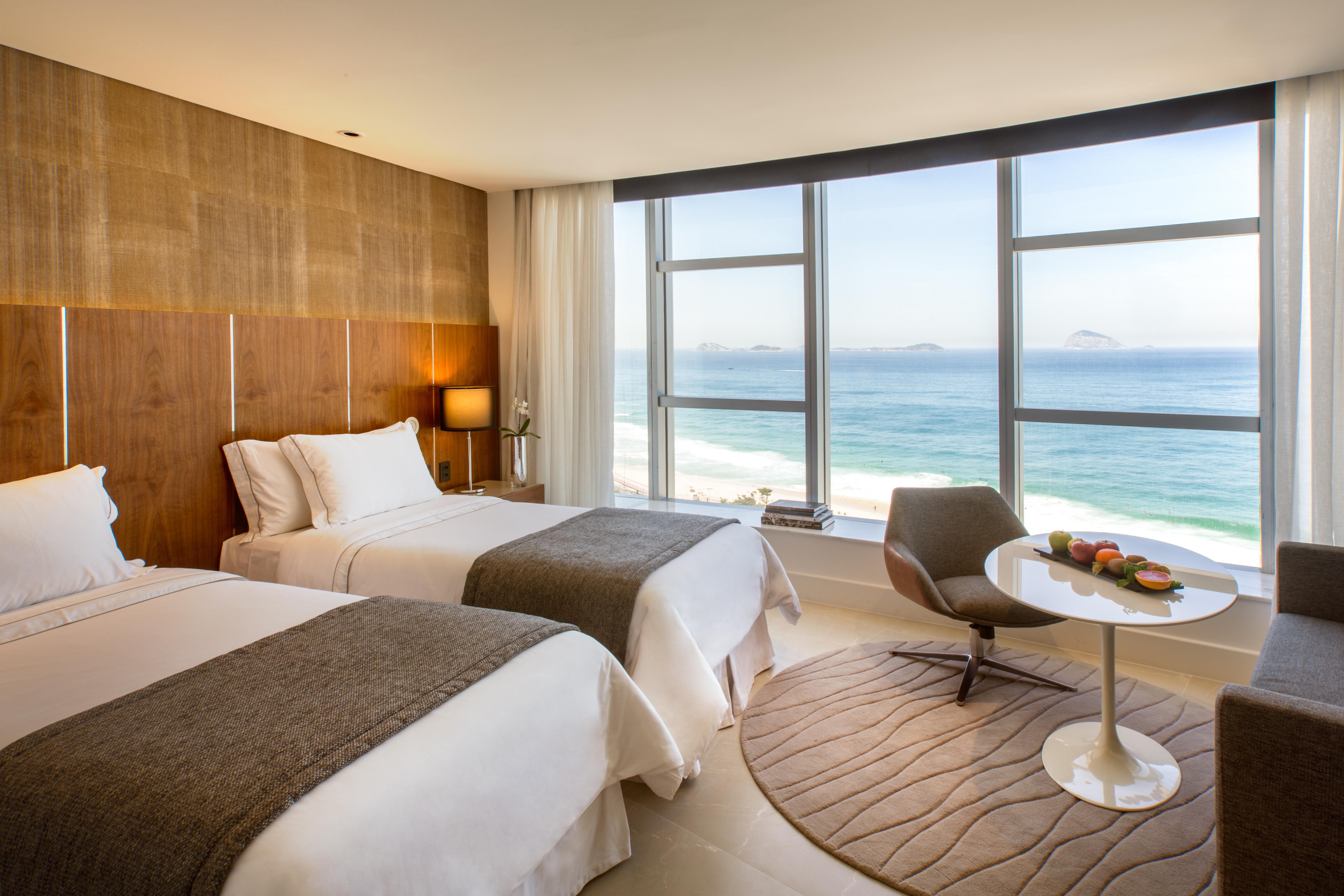 Hotel Nacional Rio De Janeiro - Oficial 外观 照片 The photo shows a modern hotel room with two beds adorned in white linens and gray throws. Large windows allow natural light to flood the space, offering a stunning view of the ocean. In the corner, there's a comfortable chair and a small round table
